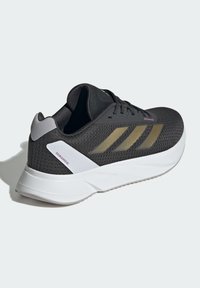 Černá síťovaná sportovní obuv se zlatými akcenty, bílou podrážkou, polstrovaným límcem a třemi paralelními pruhy. Disponuje polstrovanou mezipodrážkou pro podporu.
