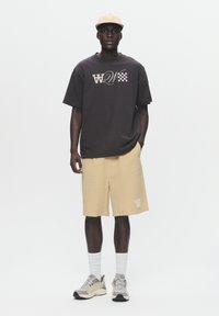 Black short-sleeve t-shirt with graphic design, paired with beige shorts. Light cap, white crew socks, and grey sneakers complete the outfit.