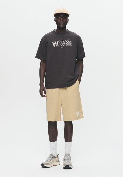 T-shirt noir à manches courtes avec un design graphique, associé à un short beige. Une casquette légère, des chaussettes blanches et des baskets grises complètent la tenue.