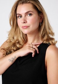 Zwart getextureerd mouwloos topje; gouden hoepel oorbellen en meerdere ringen om de vingers; licht golvend haar; helder huid met natuurlijke make-up.