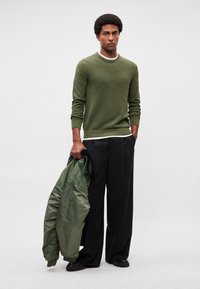 Žalios spalvos mezginys megztinis su apvalia iškirpte ir ribomis rankogaliais, derinamas su juodomis plačiąsias kelnes. Modelis laiko žalią paminkštintą striukę.