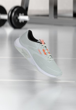 Lichtgrijze Reebok-sneaker met oranje accenten, zwevend boven een reflecterende sportschoolvloer met wazige dumbbells op de achtergrond.