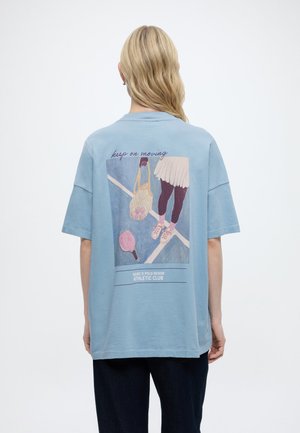 Asmuo su ilgais šviesiais plaukais dėvi šviesiai mėlyną, platų marškinėlį, ant kurio nugaros yra teniso tema iliustracija ir užrašas „keep on moving“.