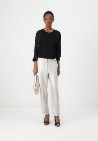Haut noir avec des manches longues plissées, associé à un pantalon large en argent brillant et des talons aiguilles noirs. Le mannequin tient une pochette beige clair.