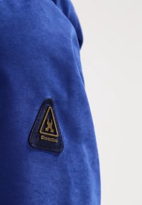 Tissu bleu à texture lisse avec un patch triangulaire noir orné de détails dorés et le mot « Gaasti » au bord inférieur.