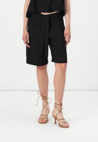 Lyhyet mustat tailored-shorts, joissa on vyöllinen vyötärö, litteät etutaskut, hieman löysä malli ja polvipituus. Tan-väriset, remmilliset korkokengät.