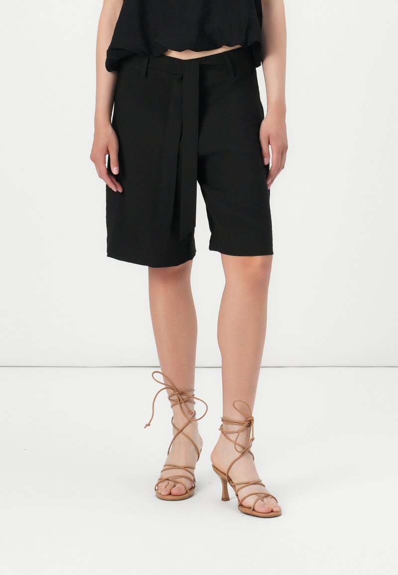 Lyhyet mustat tailored-shorts, joissa on vyöllinen vyötärö, litteät etutaskut, hieman löysä malli ja polvipituus. Tan-väriset, remmilliset korkokengät.