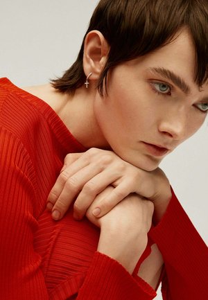 Pendientes de cruz plateados cuelgan de un peinado corto y oscuro. El sujeto lleva una camiseta de rayas rojas con textura, con las manos entrelazadas en su brazo.