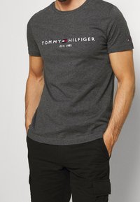 Hombre con camiseta de Tommy Hilfiger gris oscuro con logo y texto "EST. 1985", combinada con pantalones cargo negros, con las manos en los bolsillos.