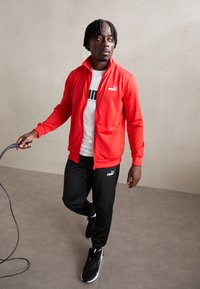 Rode zip-up jas met een hoge kraag, gecombineerd met een wit T-shirt met zwarte tekst. Zwarte sportbroeken en zwarte sneakers maken de outfit compleet.