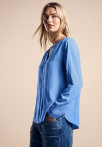 Blå blus med långa ärmar, knapplåda, mjuk textur och avslappnad passform. I kombination med denimjeans som har en lätt svans vid fållen.