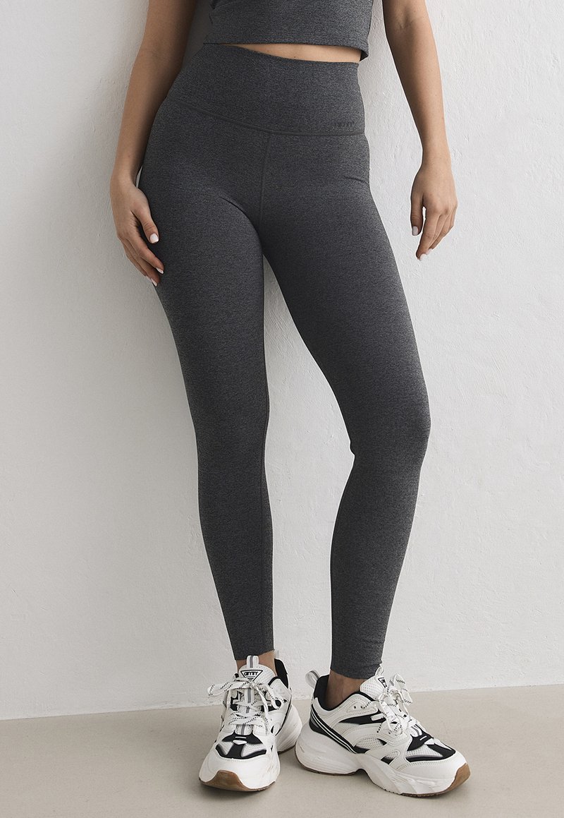 Femme portant des leggings gris foncé taille haute et des baskets blanches avec des accents noirs, debout contre un mur de couleur claire.