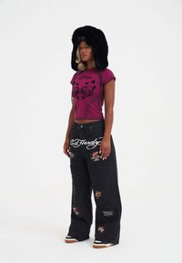 Camiseta recortada magenta con diseño gráfico negro, combinada con pantalones de mezclilla negros con parches bordados y letras blancas. Sombrero de piel negro.