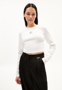 Weißes, gekürztes Langarmoberteil mit gerippter Textur und einem kleinen schwarzen rechteckigen Logo. Kombiniert mit hochgeschnittenen schwarzen Hosen.