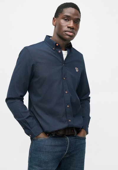 Muž s rukami vreckách, oblečený v námorníckej tmavomodrej košeli na gombíky s malou farebnou vyšívanou zebrou na hrudi a modrými džínsami.