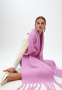 Lyserød uld tørklæde med frynser, let draperet over skuldrene. Glat tekstur, rektangulær form, båret med en let sweater og høje hæle.