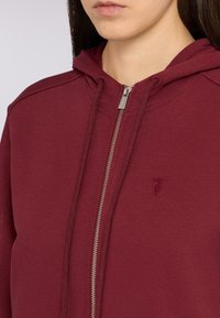 Sudadera con cremallera en burdeos, hecha de tejido suave, con un bolsillo tipo canguro, capucha con cordón y un pequeño detalle de logo en el pecho.