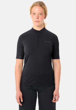 Femme à la peau claire et aux cheveux blonds, portant un maillot de cyclisme noir à manches courtes avec une fermeture éclair et un pantalon noir, debout devant un fond blanc.