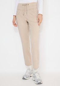 Personne portant un pantalon beige à cordon, des chaussettes blanches, des baskets argentées et un haut blanc à manches longues, debout devant un fond uni.