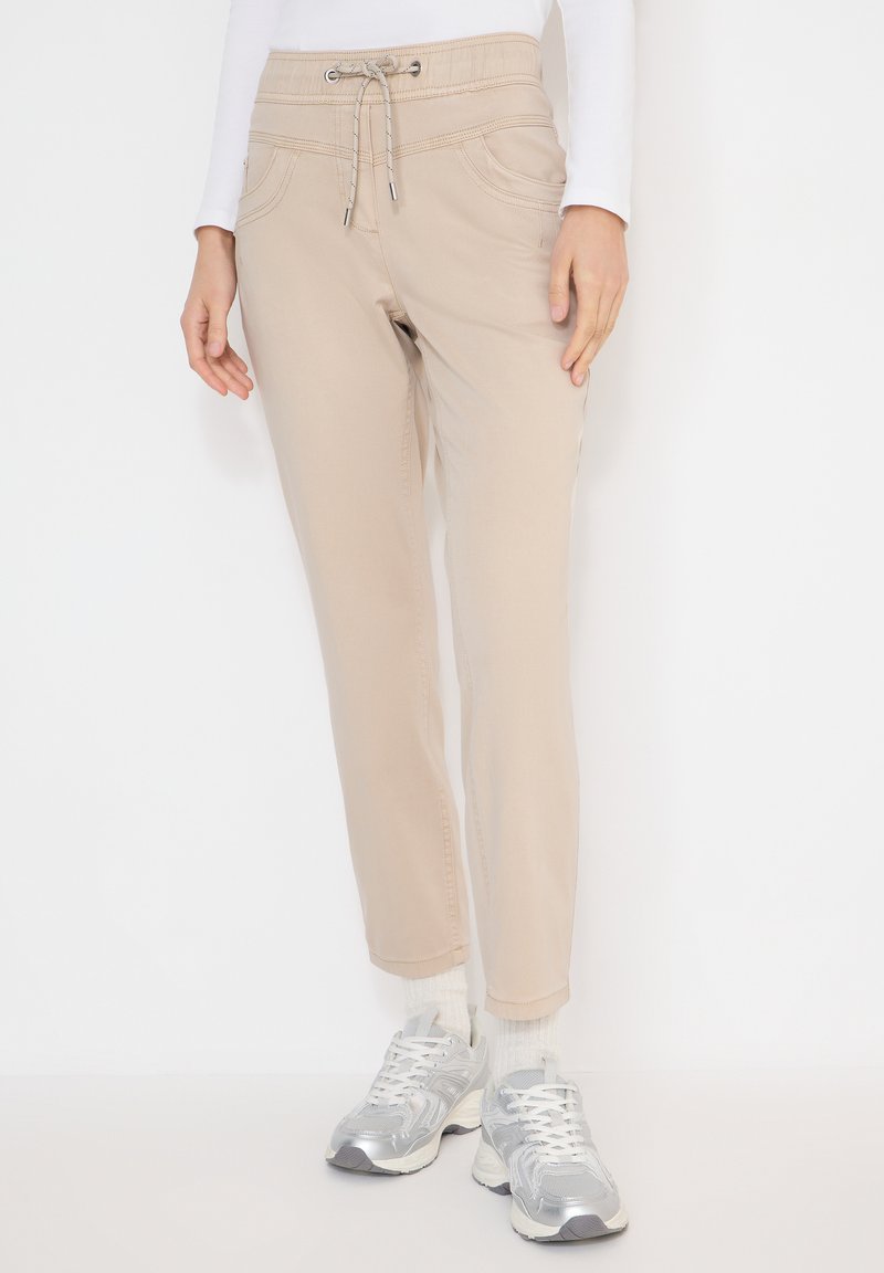 Personne portant un pantalon beige à cordon, des chaussettes blanches, des baskets argentées et un haut blanc à manches longues, debout devant un fond uni.