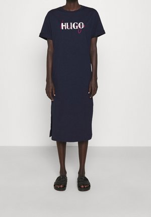 Persona que lleva un vestido midi azul marino de manga corta con aberturas laterales y texto "HUGO" en blanco y rosa en el pecho, combinado con sandalias negras.