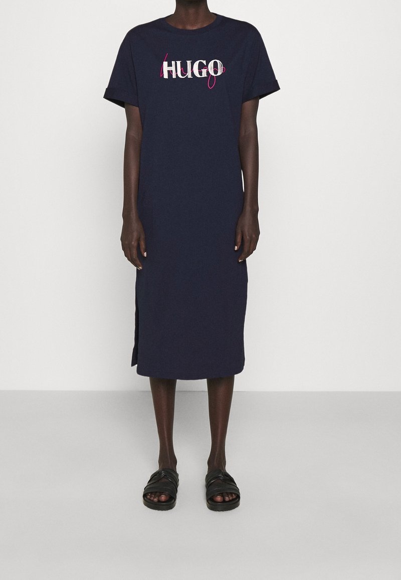 Personne portant une robe midi bleu marine à manches courtes avec des fentes latérales et un texte "HUGO" blanc et rose sur la poitrine, associée à des sandales noires.