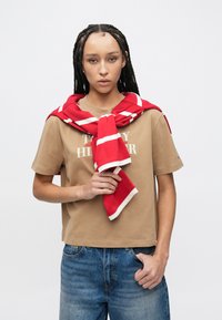 Κόκκινο-λευκό ριγέ κασκόλ δεμένο γύρω από τον λαιμό, φορεμένο πάνω από ανοιχτό καφέ μπλουζάκι. Καθημερινό μπλε τζιν με κλασική εφαρμογή.