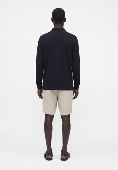 Homme debout de dos, portant une chemise noire à manches longues, un short beige et des sandales noires sur un fond blanc uni.