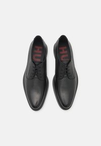 Chaussures à lacets en cuir noir avec une finition lisse et un design à bout rond. L'intérieur présente une marque "HU" rouge sur une semelle intérieure noire.