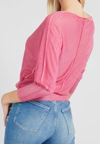 Pulóver rosa de textura suave, con mangas acanaladas y un sutil detalle de costura en la parte trasera, combinado con jeans ajustados de color azul claro.