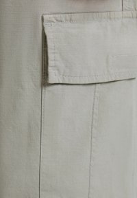 Ljust grått tyg har en framficka med lock, platta sömmar och en mjuk textur. Materialet verkar hållbart med en subtil lyster.