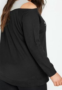 Czarna bluzka z długim rękawem i odkrytymi ramionami, wykonana z bawełny, z zaokrąglonym dołem oraz szarą warstwową dekoracją na ramionach. Z minimalną ilością tekstu.