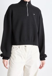Felpa nera con colletto alto, chiusura a mezza zip e orlo ed elastici sulle maniche, indossata sopra pantaloni bianchi. Presenta un piccolo logo.