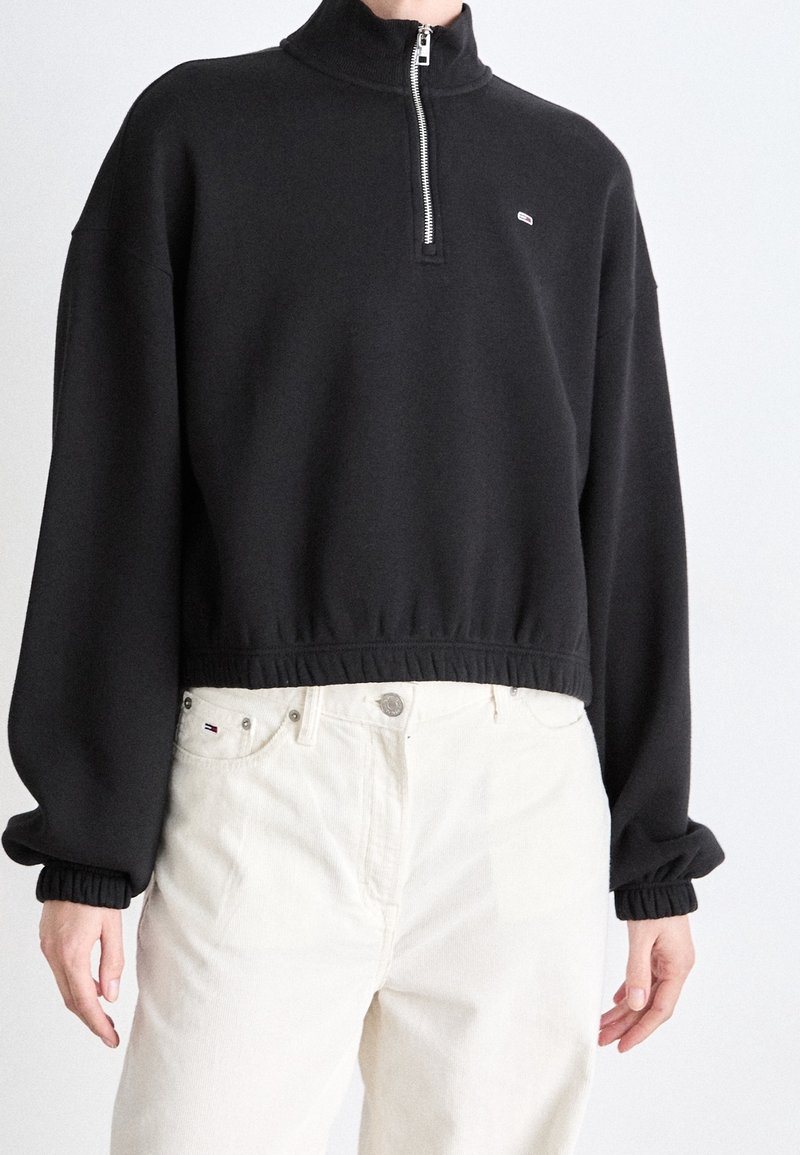 Felpa nera con colletto alto, chiusura a mezza zip e orlo ed elastici sulle maniche, indossata sopra pantaloni bianchi. Presenta un piccolo logo.