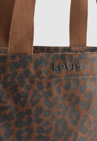 Ruda pirkinių rankinė su juodu leopardų raštu ir rudomis pintomis rankenomis, turinti mažą Levi's logotipą siuvinėtų arti viršutinio krašto.