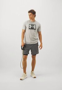Mann med lysegrå Under Armour t-skjorte, mørkegrå shorts og beige sneakers, som holder et motstandsbånd og ser bort til siden.