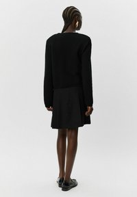 Črn pleten pulover z teksturirano površino, v kombinaciji s črno godrnjo krilo. Model nosi črne čevlje in stoji pred svetlo ozadje.