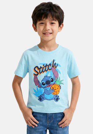 Jeune garçon souriant, portant un t-shirt bleu clair avec un personnage coloré de Stitch tenant un ananas et un jean bleu, mains dans les poches.