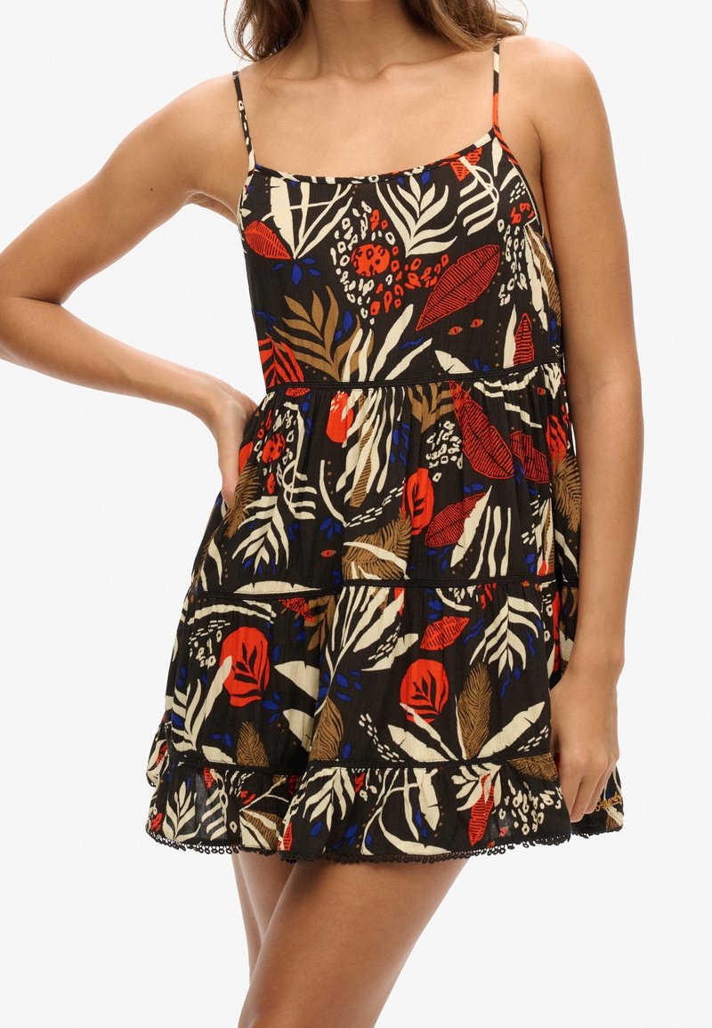 Femme portant une robe noire courte et sans manches avec un imprimé tropical feuillage et fleurs rouge, beige et bleu, une main sur la hanche.