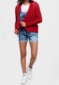 Cardigan rouge en tricot ouvert, avec un motif en losange, porté par-dessus un T-shirt blanc et un short en jean. Associé à des baskets blanches.
