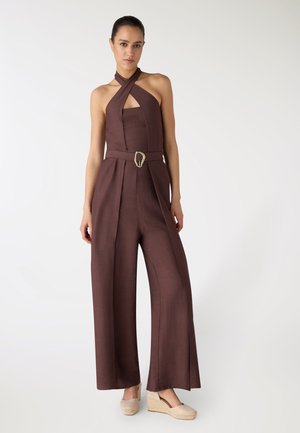 Donna che indossa una tuta senza maniche marrone con collo all'americana, gambe larghe e cintura in vita, abbinata a scarpe beige con zeppa, in piedi davanti a uno sfondo neutro.