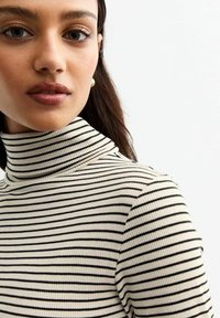 Sweter w paski z wysokim kołnierzem w kolorze kremowym i czarnym. Posiada teksturę ribbed oraz wysoki krój. Zbliżenie na materiał i dopasowanie.