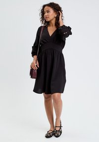 Schwarzes Kleid mit V-Ausschnitt und tailliertem Schnitt, langen Ärmeln mit Rüschensäumen. Kombiniert mit gemusterten schwarzen Ballerinas und einer burgunderfarbenen Umhängetasche.