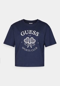 Laivansinisestä puuvillaisesta t-paidasta, jossa on lyhyet hihat, pyöreä kaula-aukko ja valkoinen "GUESS L.A. SPORTS CLUB" -grafiikka, johon sisältyy kaksi tennismailaa.