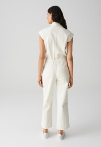 Combinaison en denim blanc avec manches courtes, taille élastique et jambes larges. Elle présente une poche arrière et un col, avec une texture lisse.