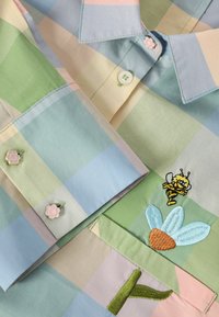 Pastelkleurig geruit overhemd met geborduurde bij- en bloemmotieven, en bloemvormige knopen op de manchetten en kraag.
