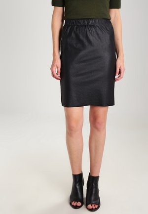 Femme portant une jupe en cuir noire mi-longue associée à des bottines noires à bout ouvert et un haut vert foncé à manches courtes.