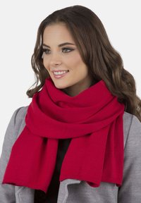Femme avec de longs cheveux bruns portant une écharpe en tricot rouge vif et un manteau gris, souriant et regardant légèrement vers la droite sur un fond blanc.