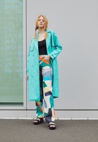 Turquoise long coat, black tank top, patterned wide-leg pants in blue, green, and orange; black platform sandals; standing against a gray backdrop.