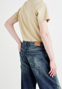 Camiseta de algodón beige combinada con jeans de mezclilla azul. Los jeans presentan un acabado texturizado, bolsillos traseros y una etiqueta de cuero marrón en la cintura.